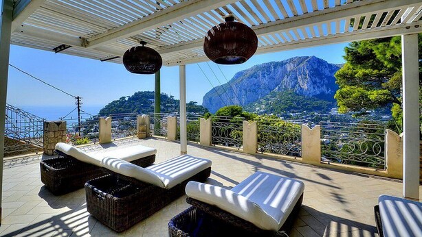 Ein überdachter Außenbereich mit Lounge-Möbeln und Korbmöbeln mit Blick auf eine Berglandschaft und das Meer.