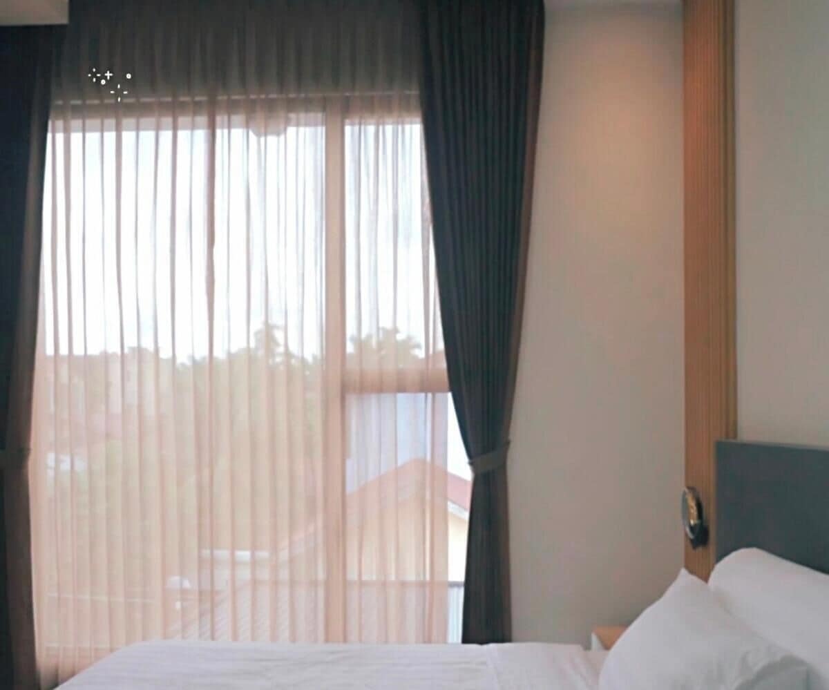 Habitación de hotel con cama, ventana con cortinas translúcidas y vistas al exterior.