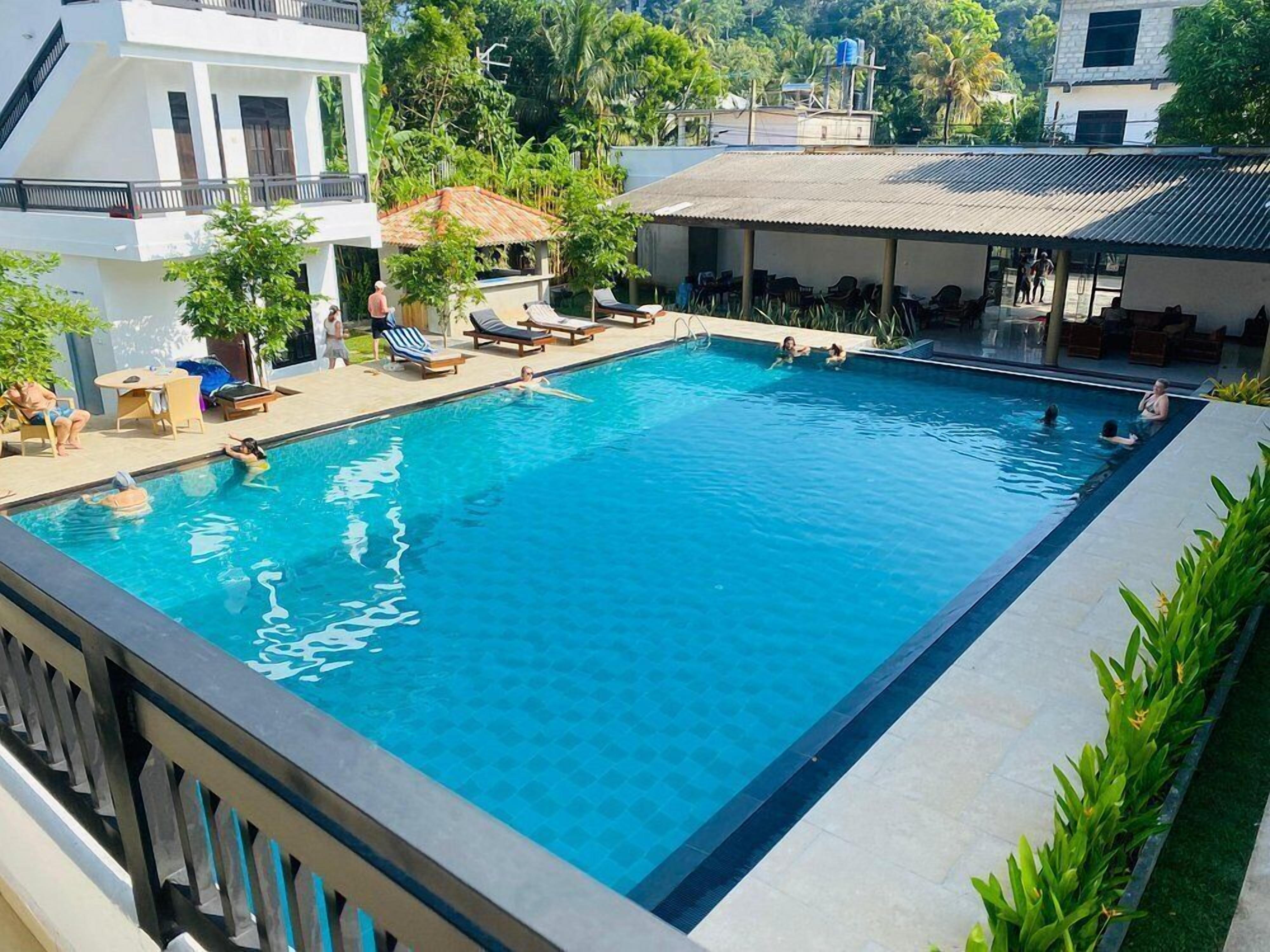 Una piscina rectangular con agua azul cristalina, rodeada de tumbonas y sombrillas, con edificios y follaje en el fondo.