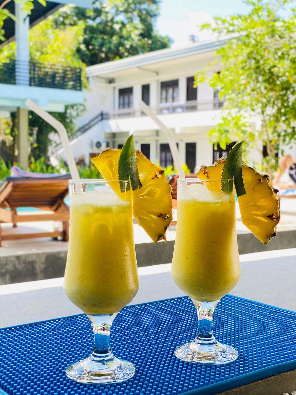Two drinks with tropical garnishes on a blue table outside a building with a pool.