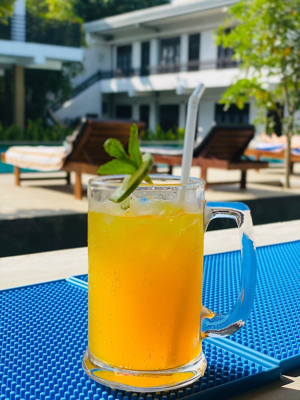 Un verre de jus d’orange, décoré d’une feuille de menthe et muni d’une paille, est posé sur une table bleue à texture particulière, près d’une piscine, avec des chaises longues et un bâtiment en arrière-plan.