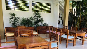 A dining area with wooden tables and chairs, large windows with blinds, and potted plants.