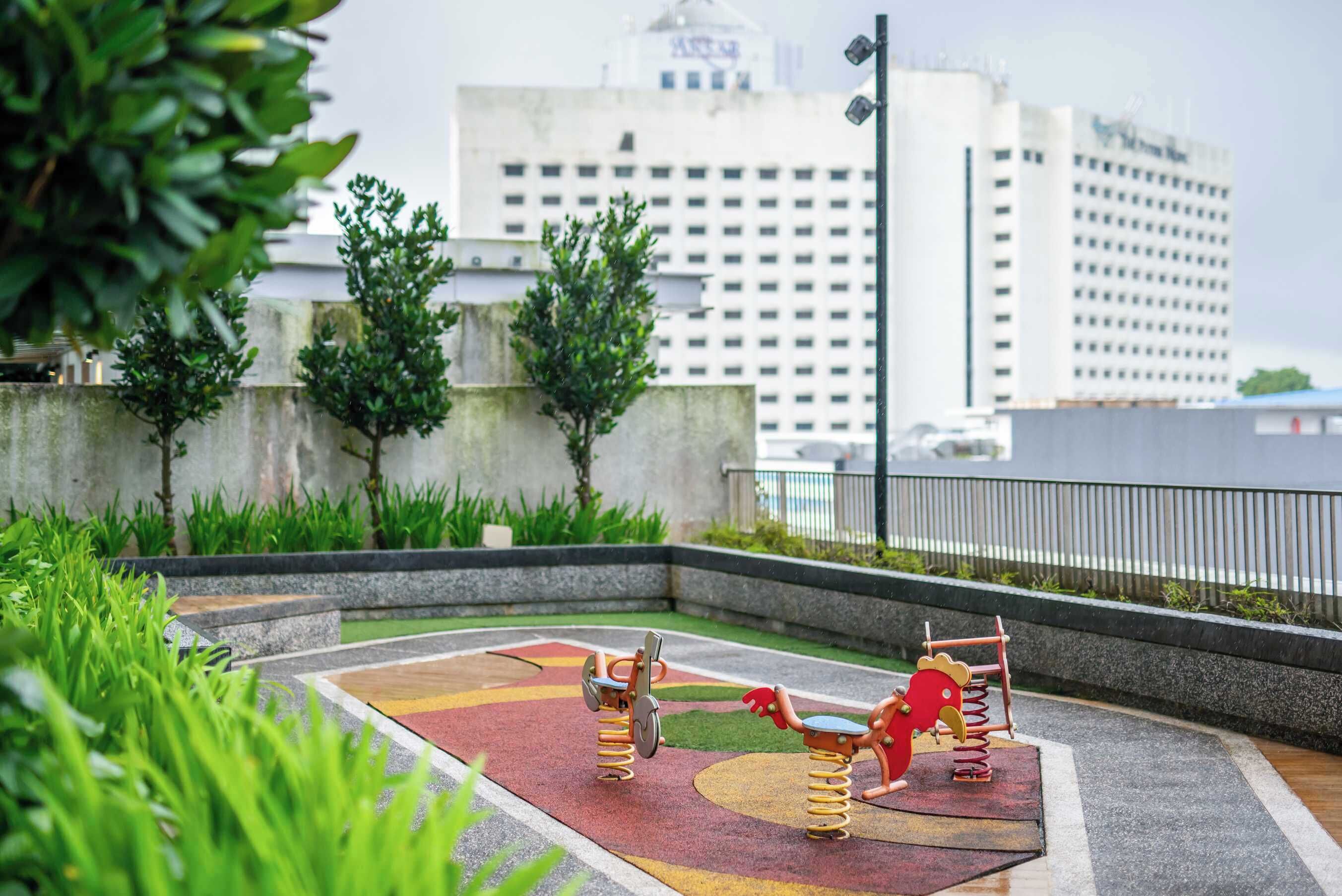A playground with two swings and a slide, surrounded by greenery and a cityscape in the background.