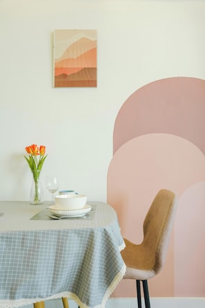 Una mesa de comedor con un florero, una copa de vino y un cuenco. Hay una silla con cojín y un cuadro colgado en la pared.