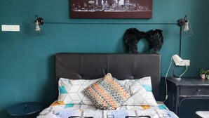 Ein Schlafzimmer mit türkisfarbenen Wänden, einem Stadtbild-Kunstwerk, einem Bett mit gemusterten Betttextilien, einem dekorativen Kopfschmuck mit Flügeln und einem Schreibtisch mit Lampe.