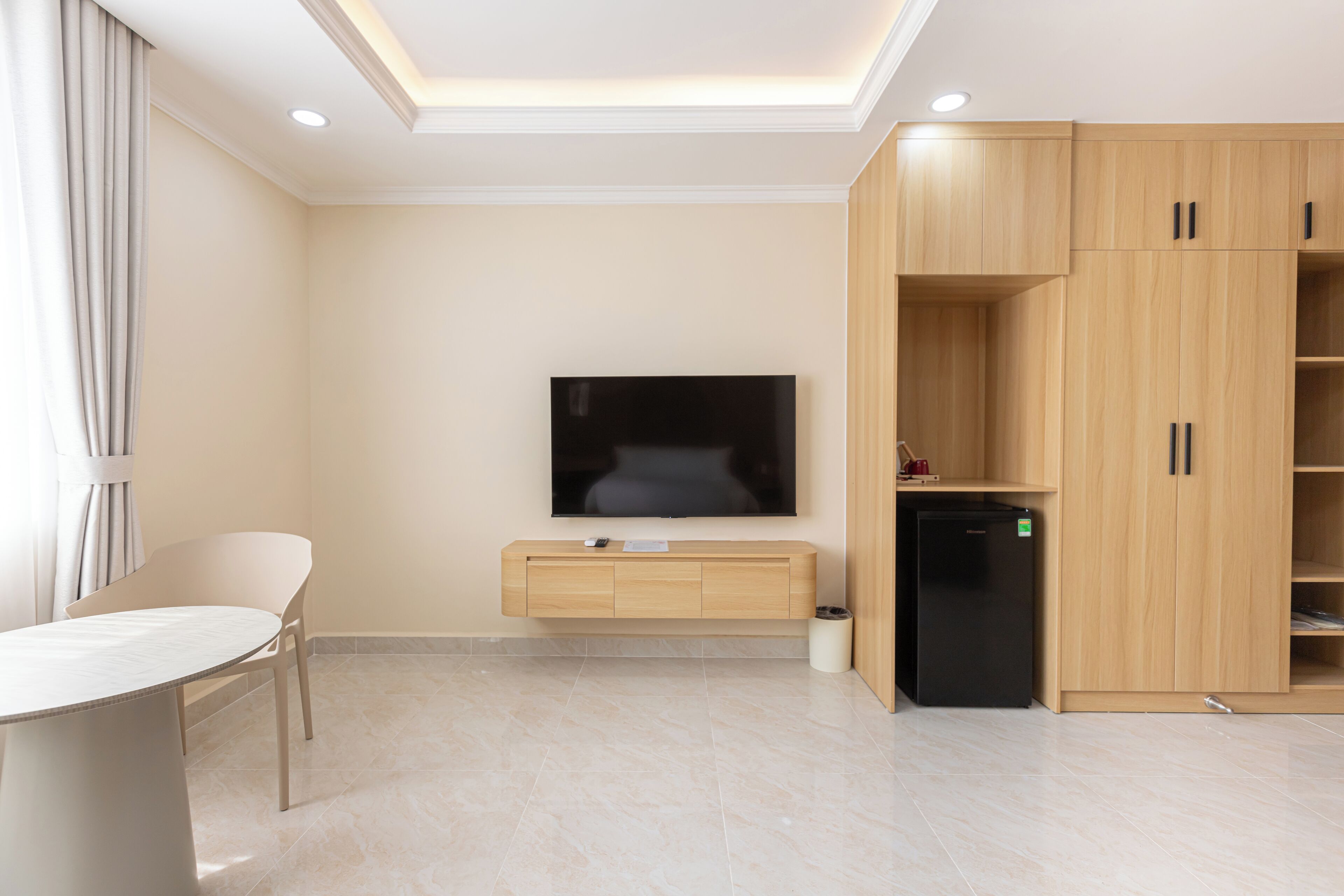 A modern living room with a flat-screen TV, wooden cabinets, a small black refrigerator, and a round table with a chair.