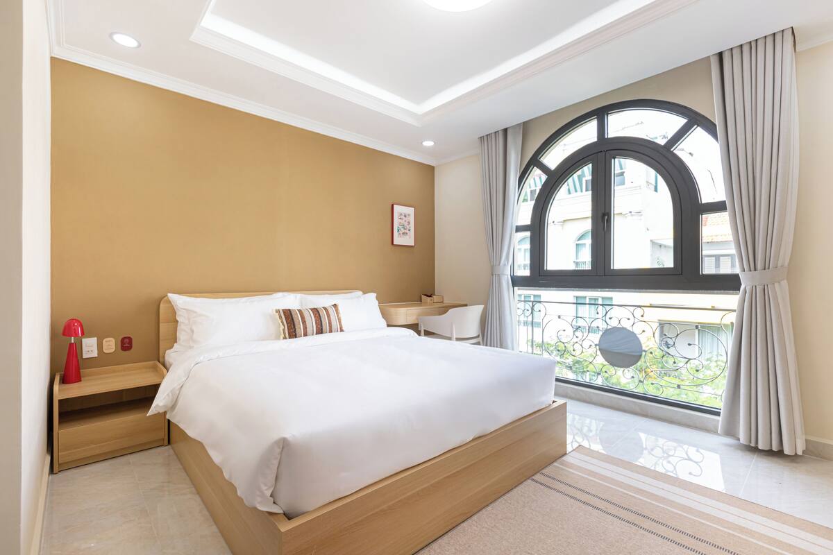 Un dormitorio con una cama grande, una mesita de noche, cortinas en la ventana y vistas a edificios.