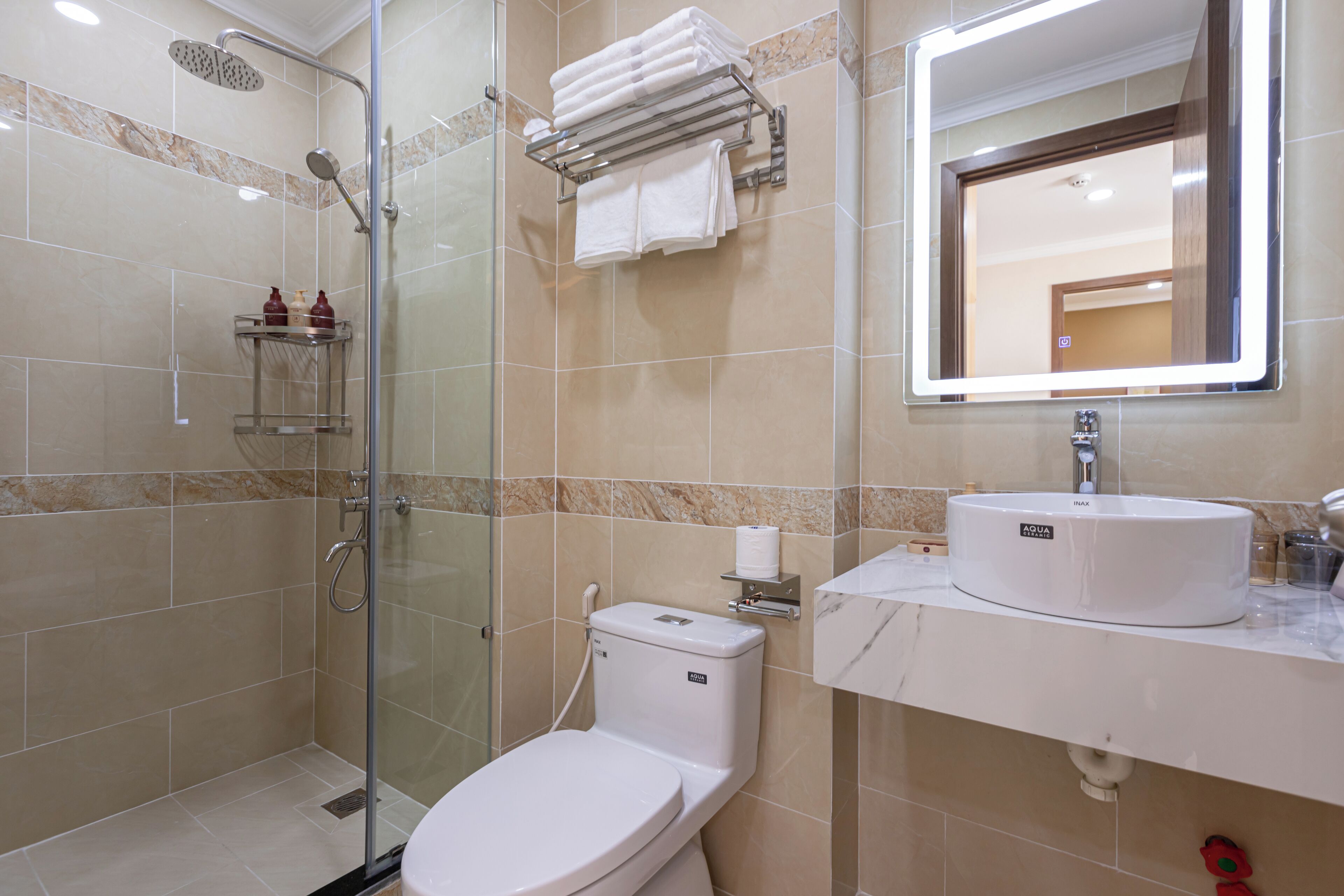 A modern bathroom with a glass-enclosed shower, a white toilet, a sink with a marble countertop, and a mirror with a lighted frame.