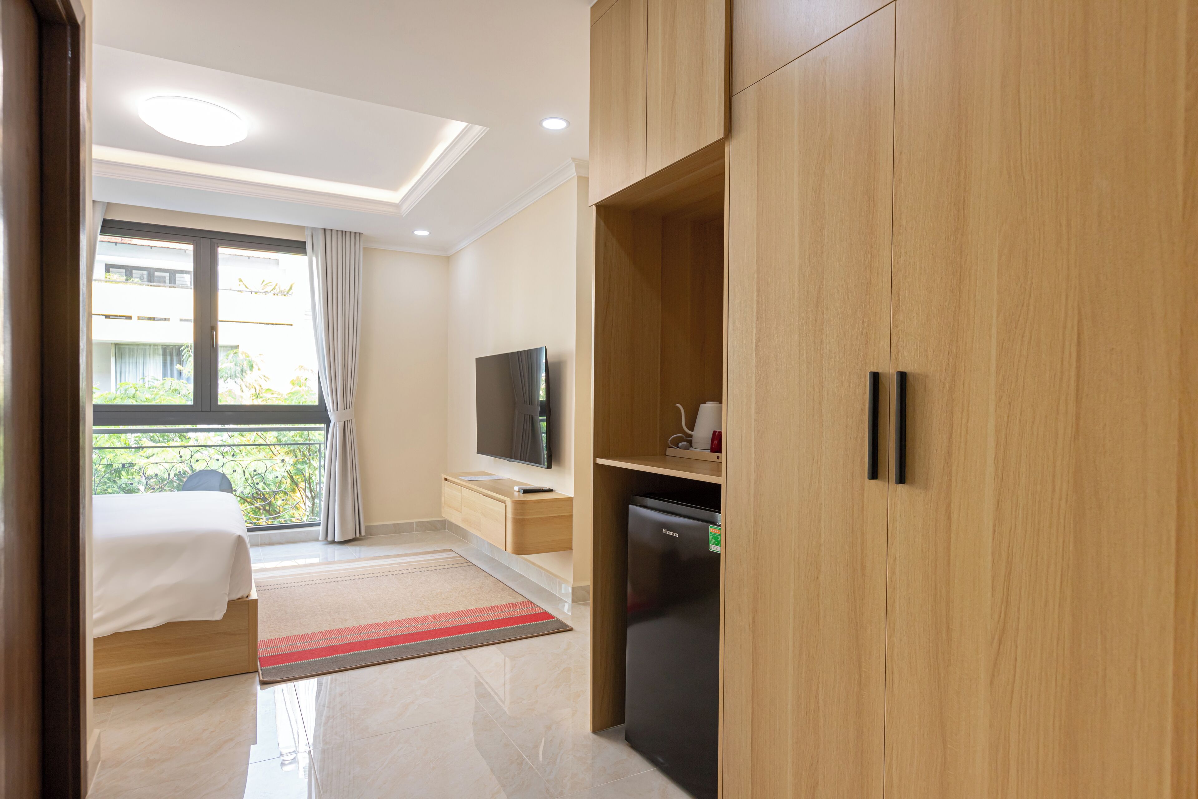 A hotel room with a bed, a TV, a refrigerator, wooden cabinets, and a balcony with a view of greenery.