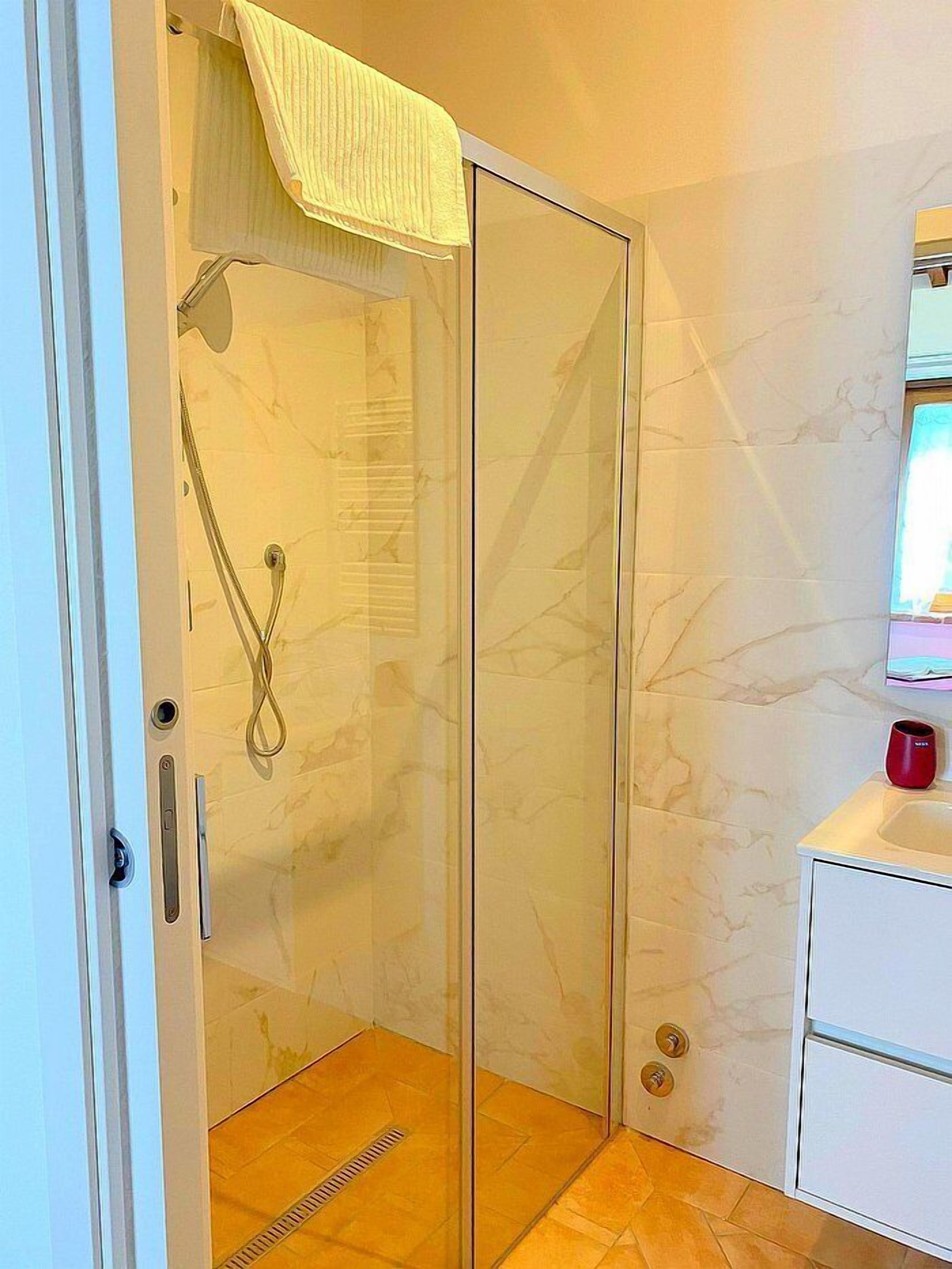 Une douche fermée par des parois de verre, avec une serviette suspendue au-dessus, un lavabo blanc et une tasse rouge, ainsi qu’un mur à motifs de marbre.