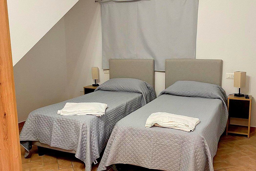 Een kamer met twee eenpersoonsbedden, elk voorzien van nachtkastje en lamp. De kamer heeft een schuine kap en een gordijn.