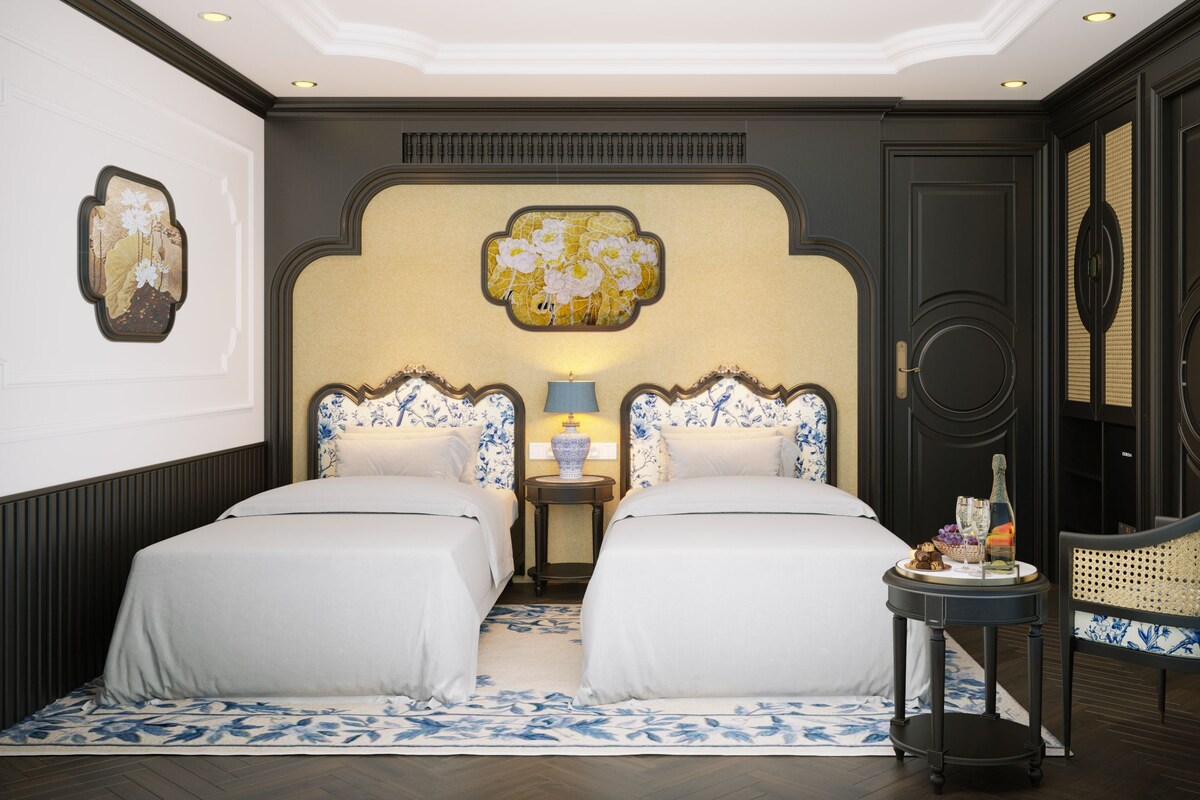 A hotel room with two beds, a small table with a lamp, a decorative wall piece, and a wicker chair.