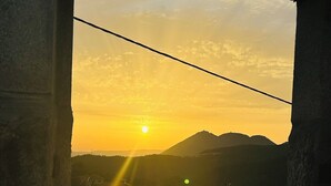 Une vue sur le coucher de soleil à travers une fenêtre ornée d’un cadre en pierre, avec une montagne visible au loin et un bâtiment doté d’un toit rouge.