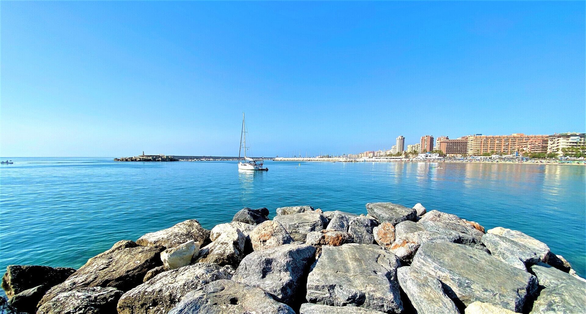 A coastal scene with a rocky breakwater, calm blue waters, and a sailboat.