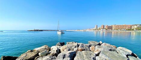 A coastal scene with a rocky breakwater, calm blue waters, and a sailboat.