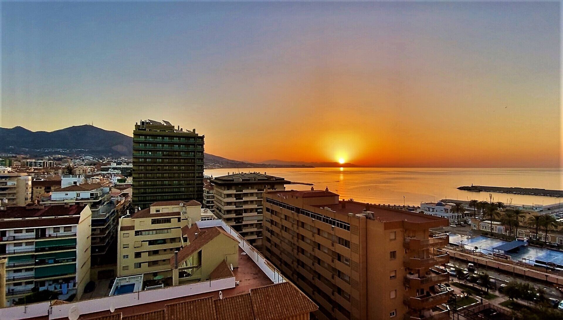 Une vue urbaine avec un coucher de soleil sur une étendue d’eau, mettant en scène des bâtiments modernes et un gratte-ciel imposant.
