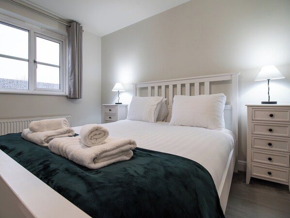 A neatly made bed with white linens and a green blanket, a bedside table with a lamp, and a window with curtains. - 4-Bed Home near Telford Centre (Telford and Wrekin)