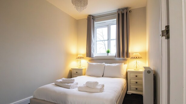 Ein ordentlich bezogenes Bett mit weißen Leinentüchern und Handtüchern, zwei Nachttische mit Lampen, ein Fenster mit Vorhängen und eine Heizung.