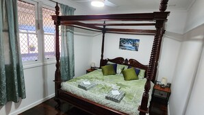 Ein Bett mit Baldachin, dessen Holzbettgestell einen grünen Überwurf und zahlreiche Kissen trägt. Über dem Bett befindet sich ein Deckenventilator, und zur Linke eine blickdichte Gardine am Fenster.