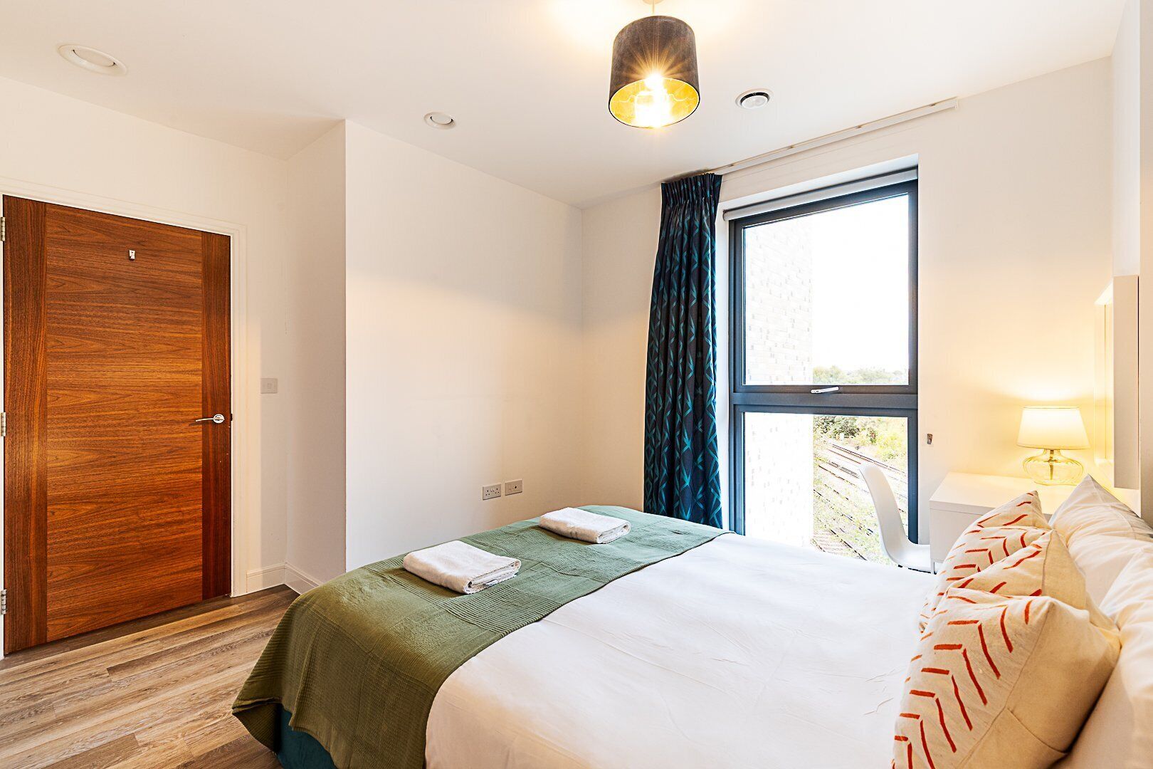 A hotel room with a wooden door, a bed with a green bedspread, a white pillow, a window with a view of a building, and a nightstand with a lamp.
