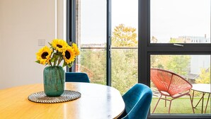 Une pièce moderne avec une table ronde en bois, un vase vert contenant des tournesols, une chaise bleue et une chaise rouge donnant sur un paysage urbain grâce à de grandes fenêtres.
