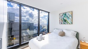 Une chambre d’hôtel moderne avec un grand lit, du linge de lit blanc, une vue sur la ville grâce à de grandes fenêtres du sol au plafond et une œuvre d’art abstraite accrochée au mur.