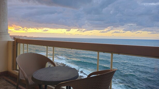 A balcony with wicker chairs and a small table, overlooking a body of water with a distant city skyline during sunset. - Midpoint sea view - families only (Alexandria)