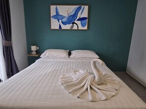 Un lit avec une couette blanche et une serviette décorative en forme de cygne, un tableau représentant des formes abstraites bleues accroché au mur, et une table de chevet avec une lampe.