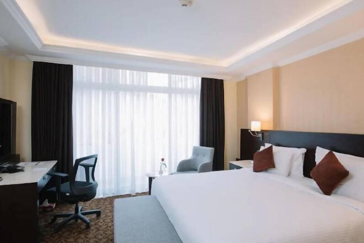 A hotel room with a large bed, a desk, a chair, a small table, and a window with curtains.