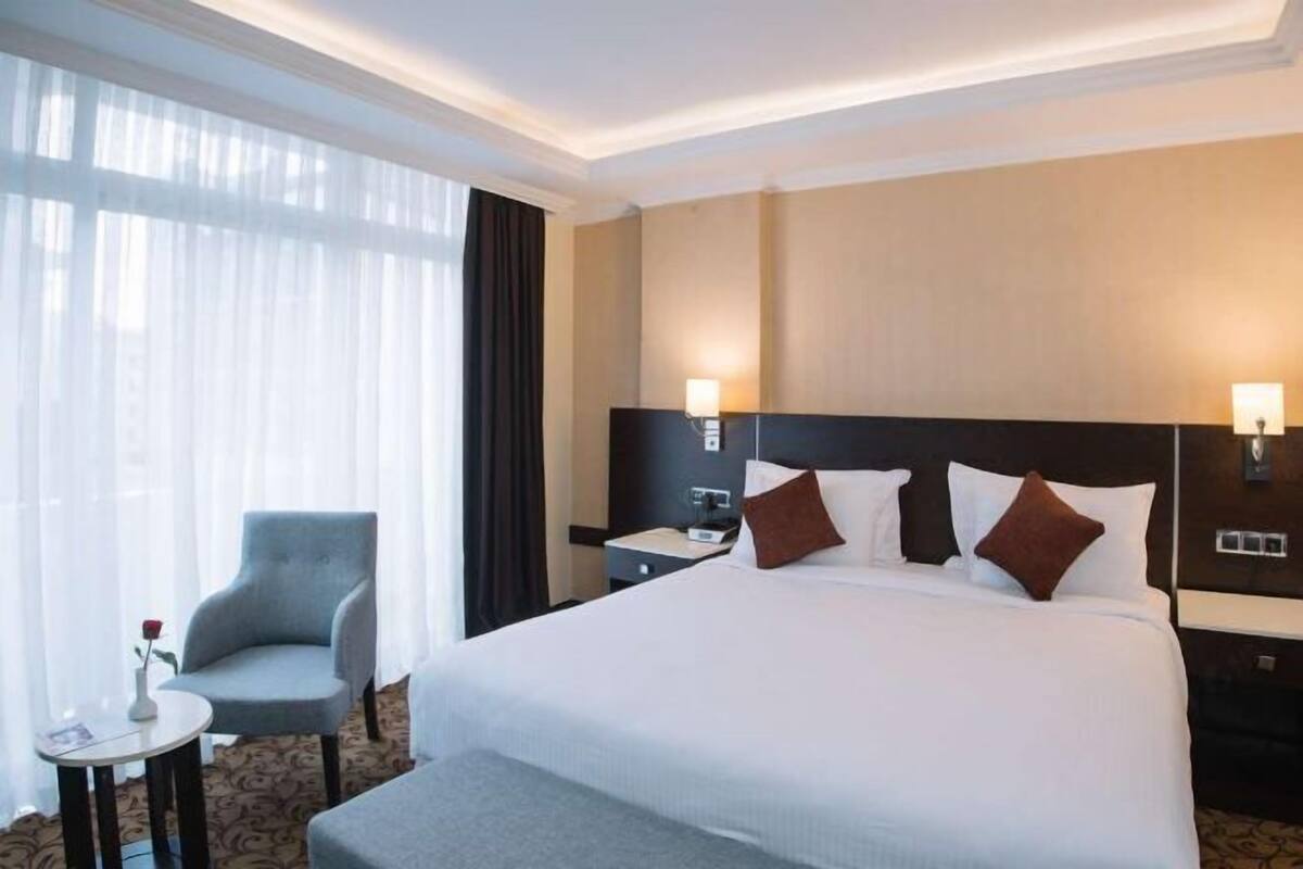 A hotel room with a large bed, a bedside table, a chair, a small table with a vase, and a window with curtains.