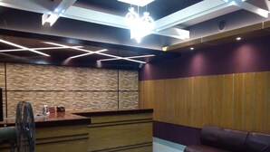A reception area with a wooden reception desk, a seating area with a leather couch, and a wall with a geometric light fixture. - Hotel Marine Plaza (Cox's Bazar)