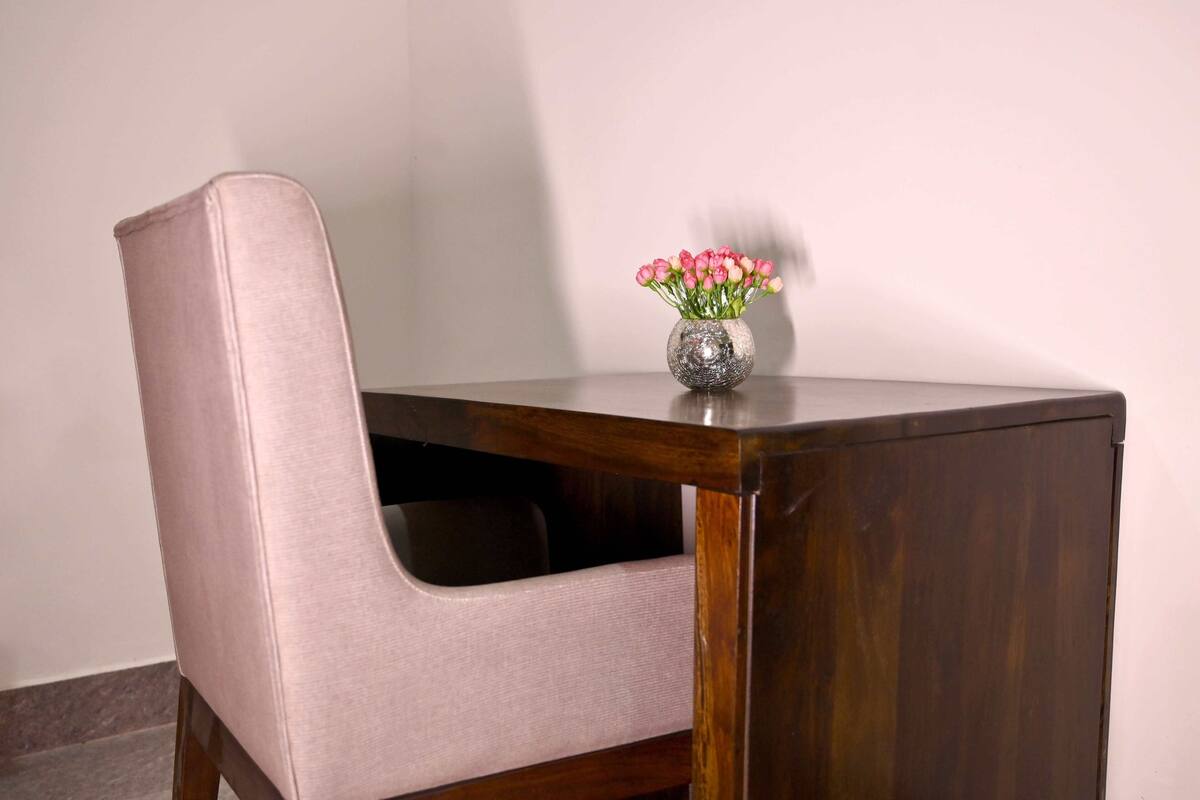 Une chaise dotée d’un dossier rembourré et d’un petit vase rond en métal, contenant des fleurs roses, posé sur une table en bois.