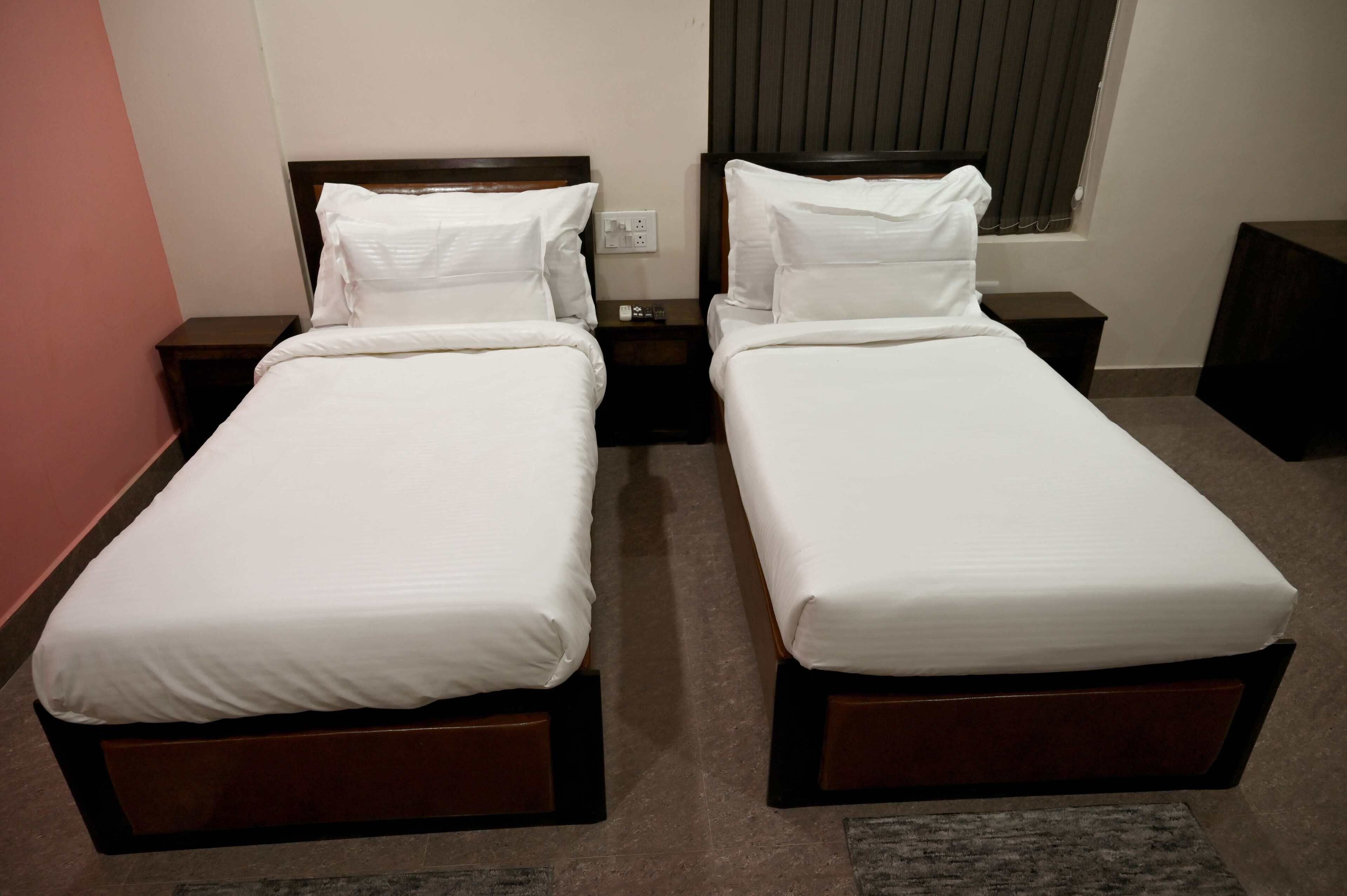 Een hotelkamer met twee enkele bedden, houten hoofdborden, een klein nachtkastje met een lamp en een raam met zonwering.