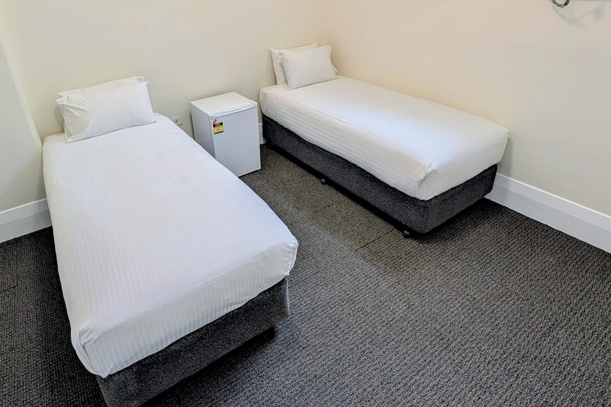 A small, clean room with two single beds, a small white bedside table, and a compact refrigerator.
