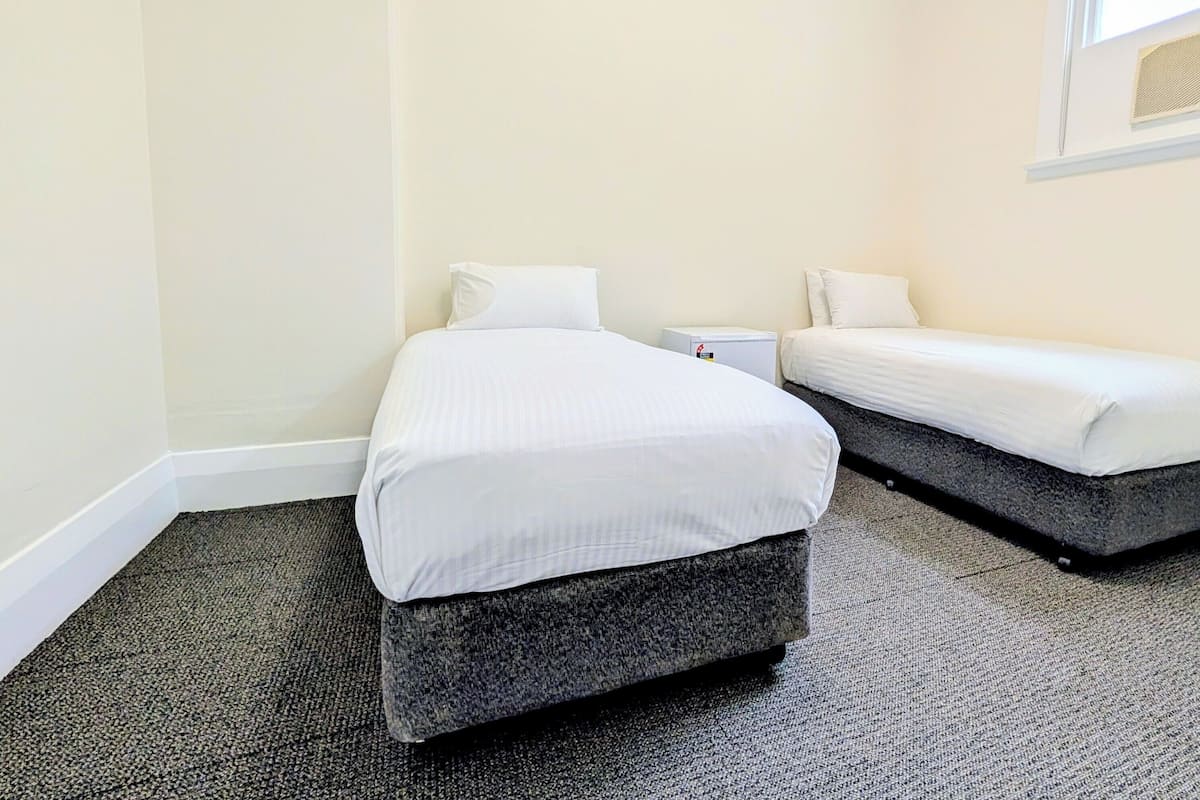 A small, clean room with two single beds, a small bedside table, and a wall-mounted heater.