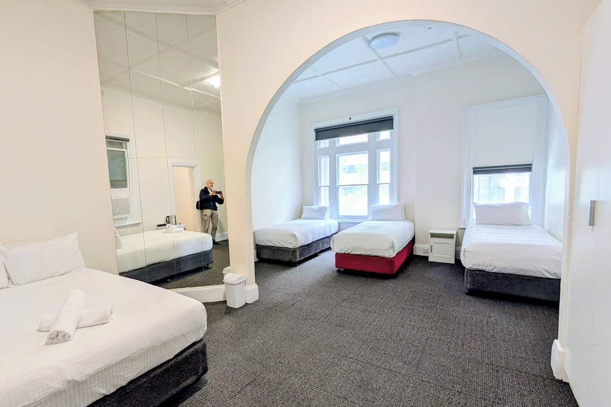 A hotel room with three beds, a large arched window, and a mirror reflecting the room.