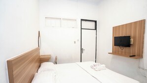 A bedroom with a wooden headboard, a bed with white bedding, a small bedside table, a TV mounted on a wooden panel, and a door with a black frame. - Tamastu House Pasar Baru Jakarta (Jakarta)