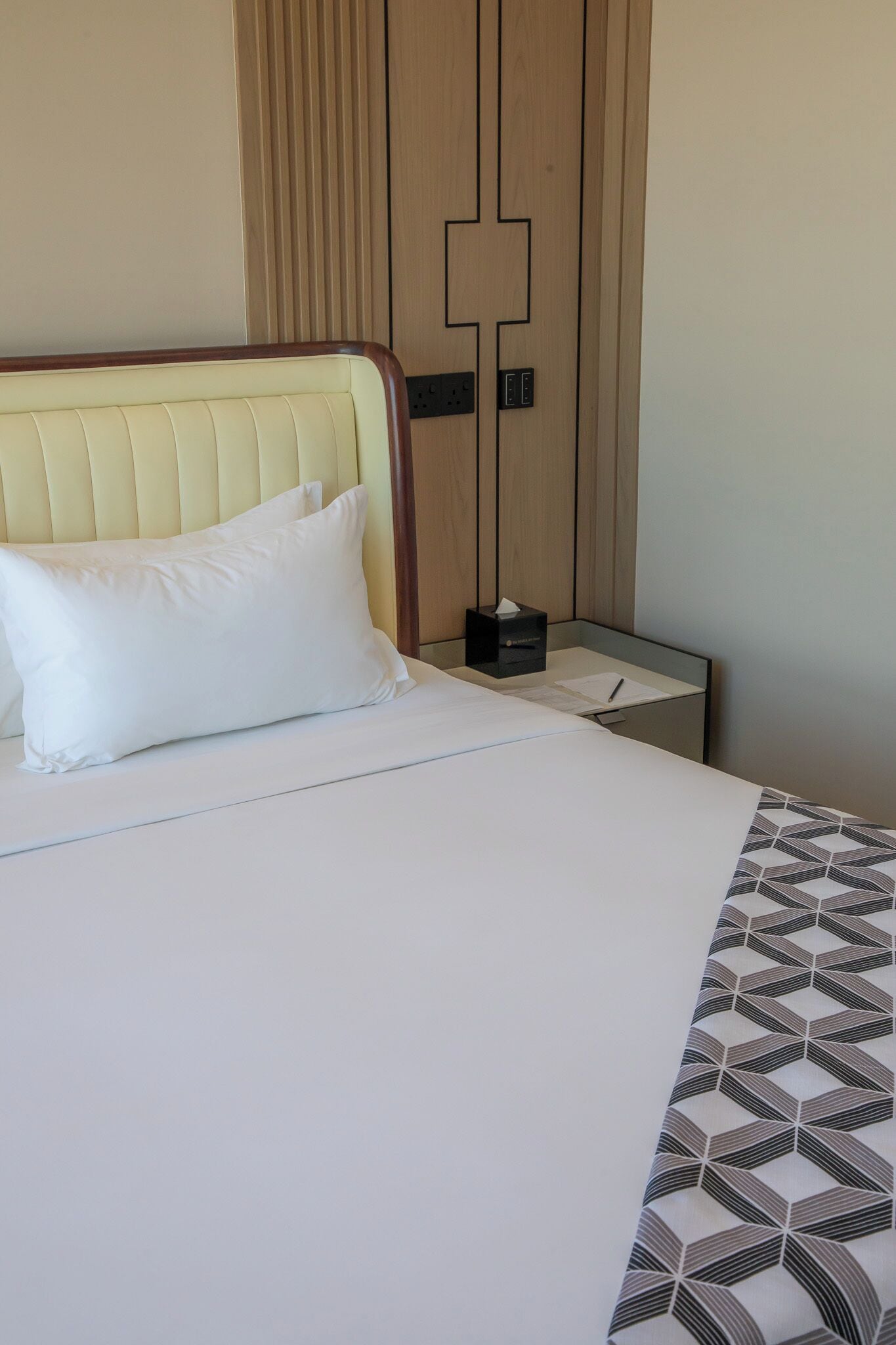 A hotel room with a bed, a bedside table with a tissue box, and a wall with a geometric pattern.