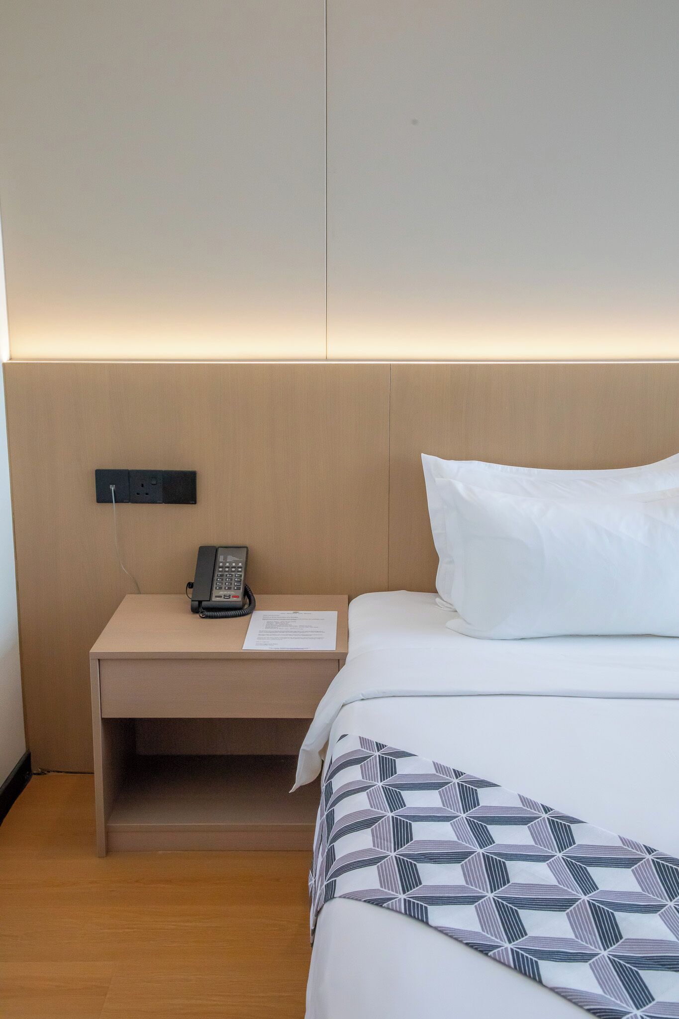 A hotel room with a bed, a bedside table with a phone, and a small shelf.