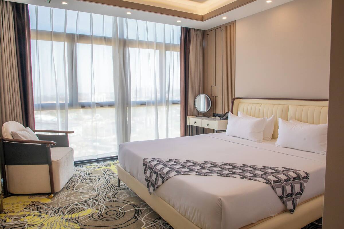 Ein Hotelzimmer mit Bett, Sessel, Nachttisch mit Spiegel und Blick durch große Fenster.