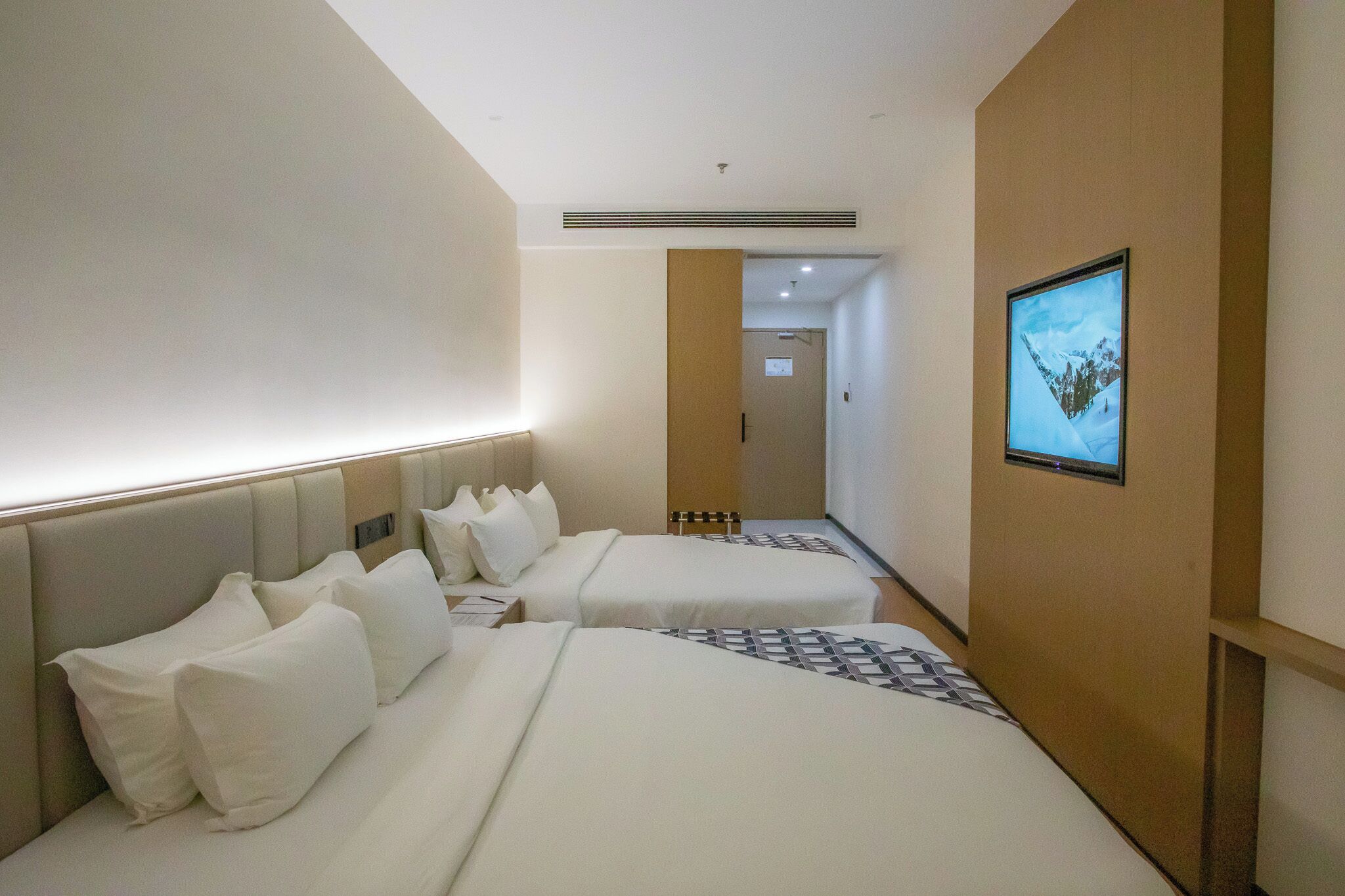 A hotel room with two beds, a TV mounted on the wall, and a hallway leading to another room.