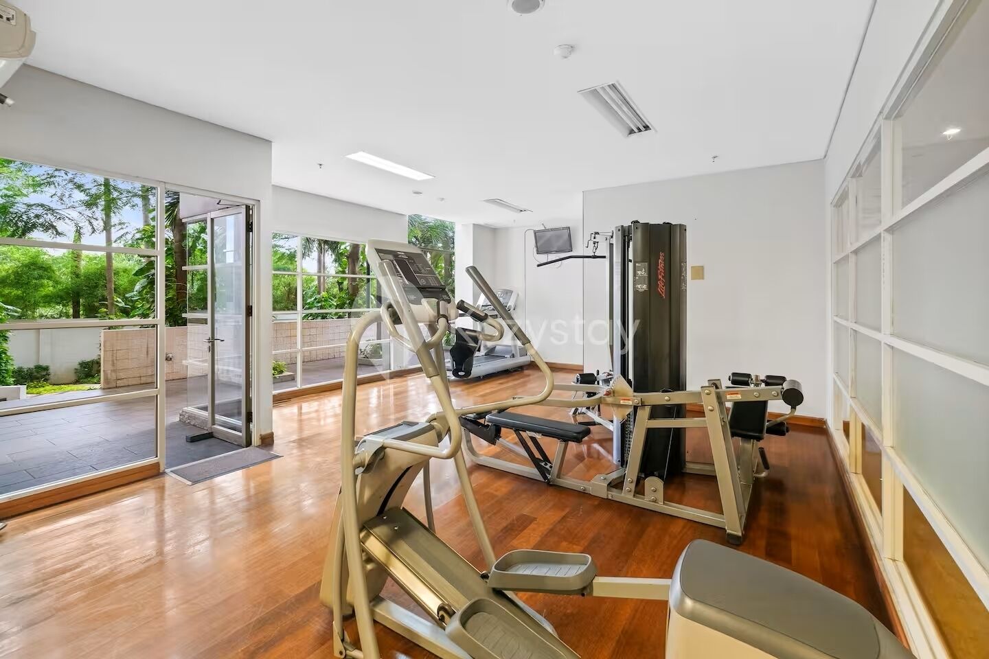 A modern gym with exercise equipment, large windows, and a view of greenery.
