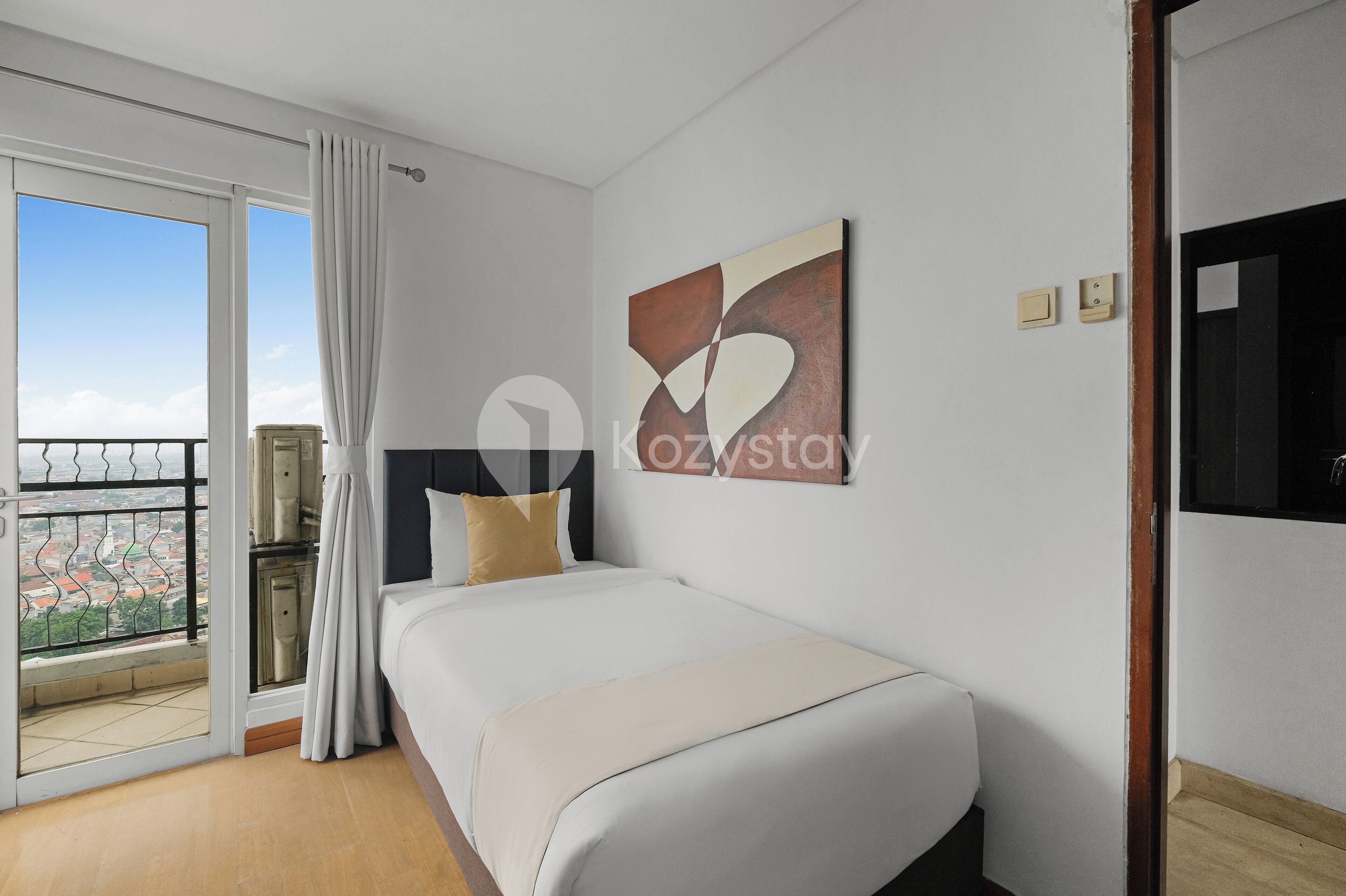 Habitación de hotel con una cama, vista de la ciudad desde un balcón y un cuadro abstracto moderno en la pared.