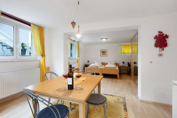 Un coin repas avec une table en bois, des chaises et une bouteille de vin. On aperçoit un lit et un bureau à l’arrière-plan.