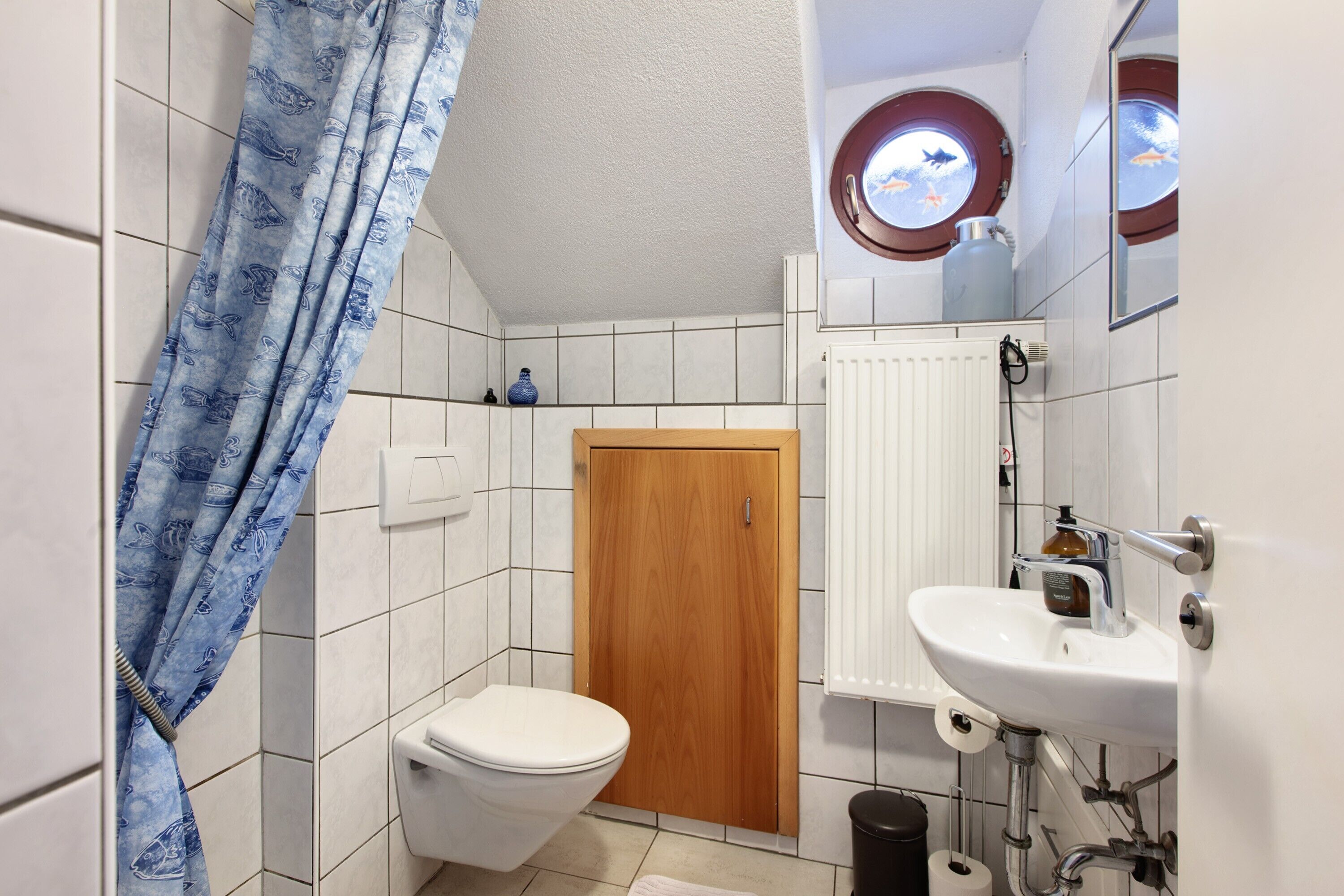 Une salle de bain avec des toilettes, une porte en bois, un lavabo blanc, une fenêtre donnant sur un aquarium et un rideau de douche bleu.