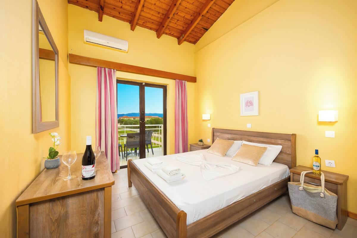 A bedroom with a wooden bed, a nightstand with a bottle, a bedside table with a wine bottle, a mirror, and a view of the outdoors through a glass door.