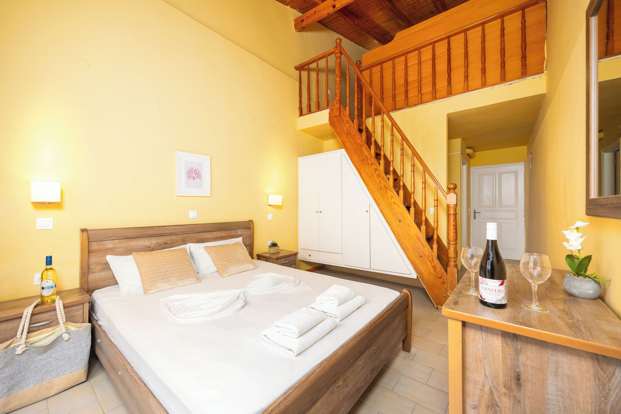 A hotel room with a wooden bed, a white wardrobe, a wooden staircase, and a small table with a bottle and glasses.