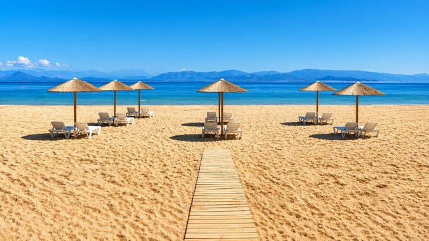 A beach with sun loungers and thatched umbrellas, a wooden walkway, and a clear view of the sea and mountains in the background.