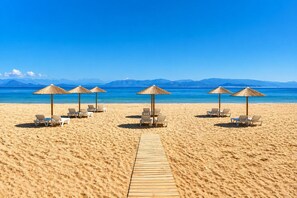 A beach with sun loungers and thatched umbrellas, a wooden walkway, and a clear view of the sea and mountains in the background. - Koulouris Beach Hotel (Kavos)