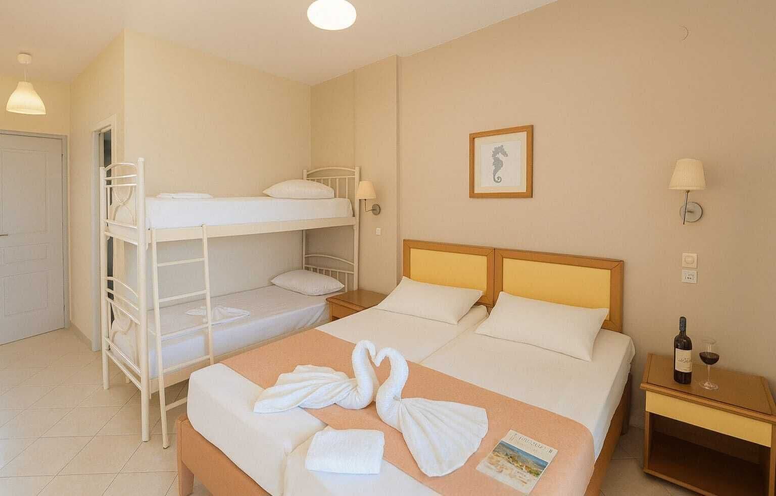 A hotel room with bunk beds, a bed with towels, a framed picture, a bottle of wine, and a nightstand.
